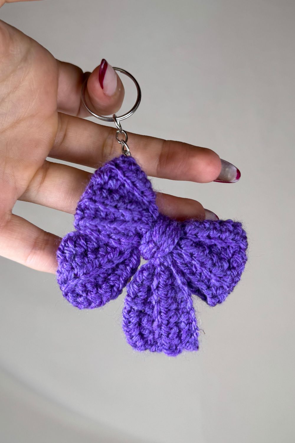 Portachiavi a fiocco viola realizzato all’uncinetto, fatto a mano in Italia da Made By Hand, mostrato in mano per evidenziare i dettagli artigianali e la dimensione reale