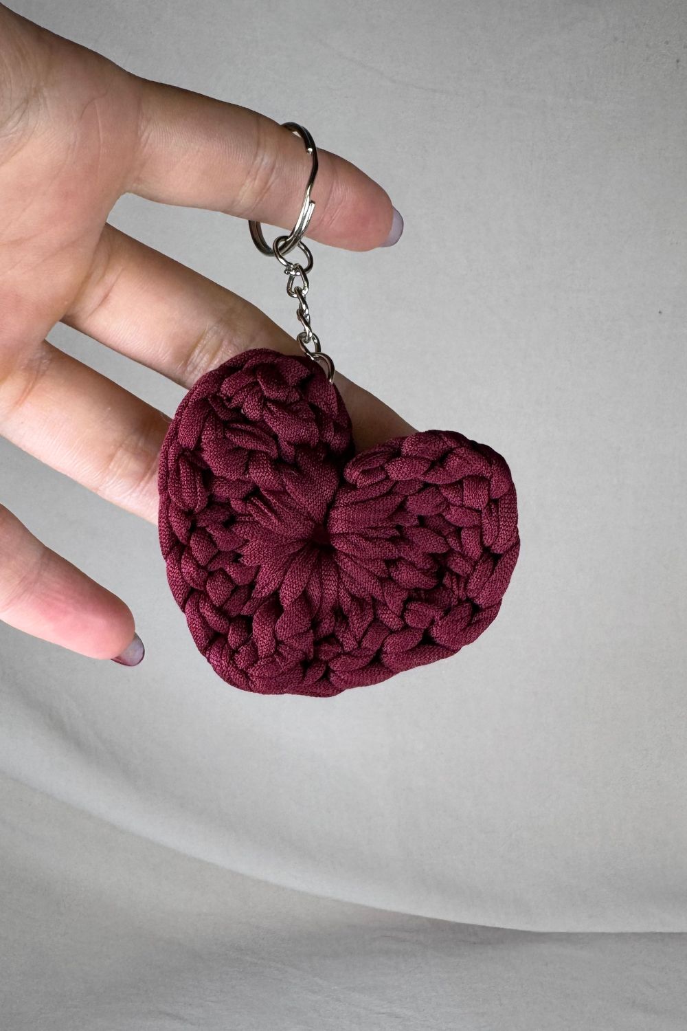 Portachiavi cuore bordeaux lavorato all’uncinetto con fettuccia, realizzato a mano in Italia da Made By Hand, mostrato in mano per dare un’idea delle dimensioni