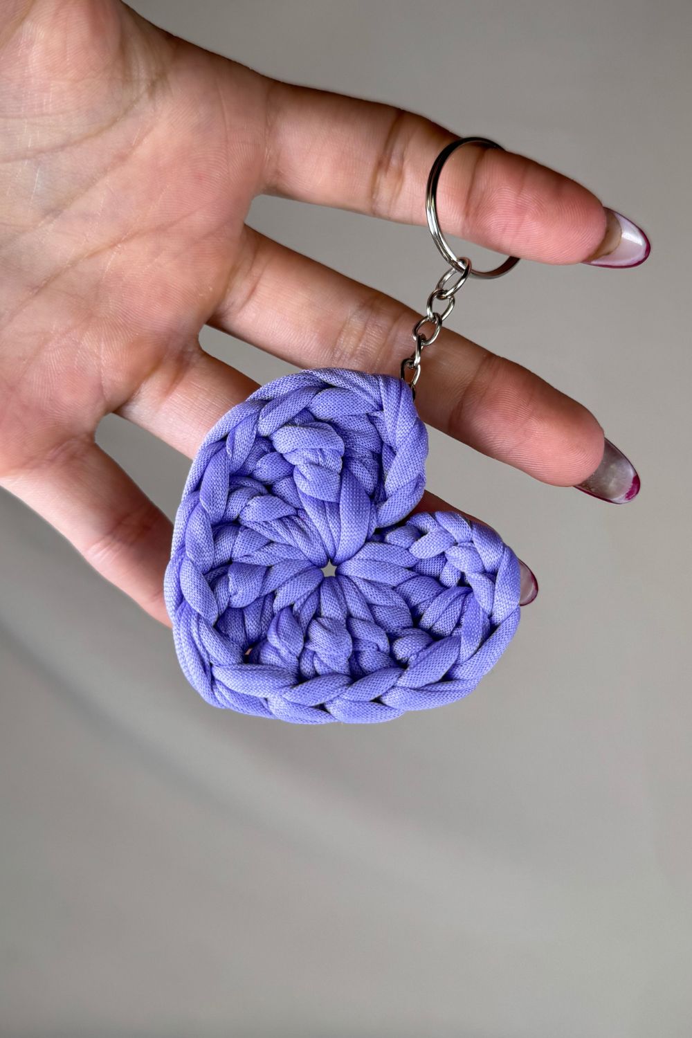 Portachiavi a cuore lilla realizzato all’uncinetto con fettuccia, fatto a mano in Italia da Made By Hand, mostrato in mano per dare un’idea reale delle dimensioni