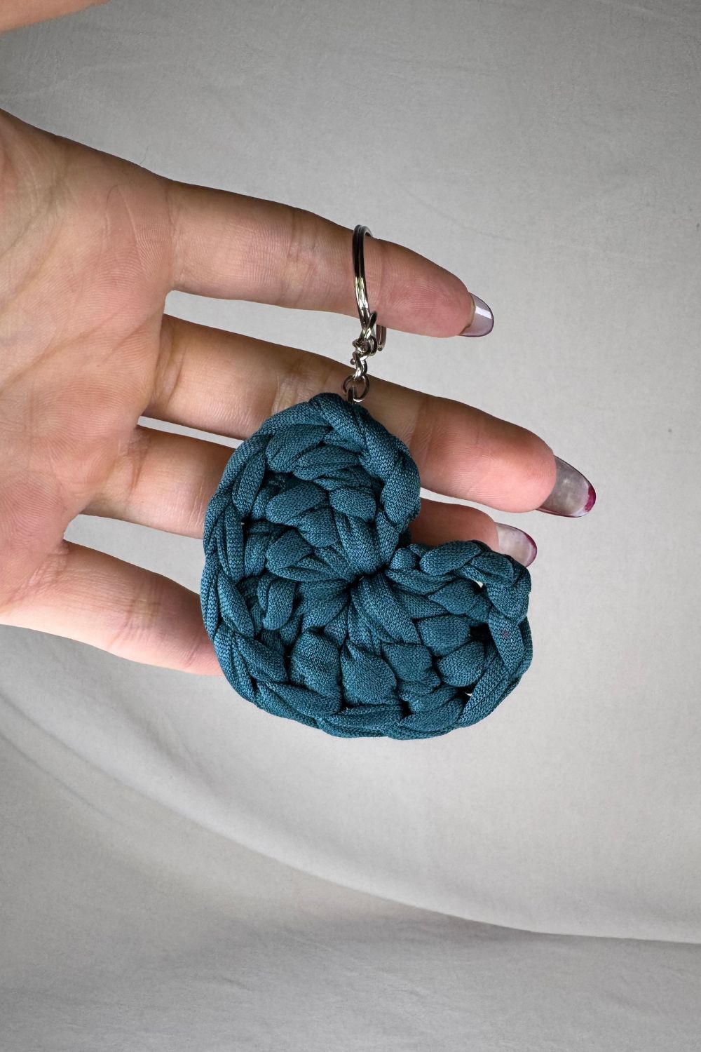 Portachiavi a cuore color petrolio lavorato all’uncinetto con fettuccia, fatto a mano in Italia da Made By Hand, mostrato in mano per evidenziare i dettagli artigianali