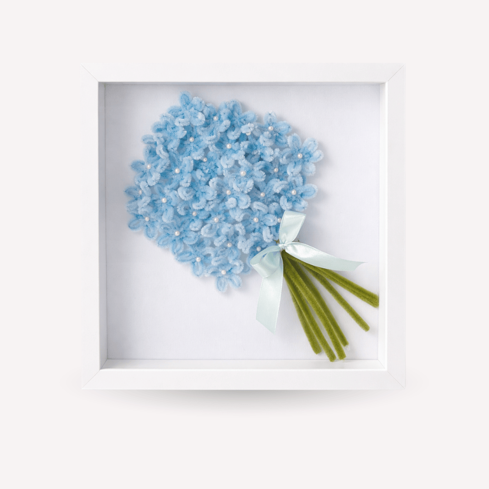 Quadro ricordo artigianale con cornice bianca, bouquet di fiori azzurri fatti a mano con piccoli dettagli luminosi, fiocco celestino e steli verdi. Decorazione elegante e personalizzabile, adatta come idea regalo o complemento d’arredo