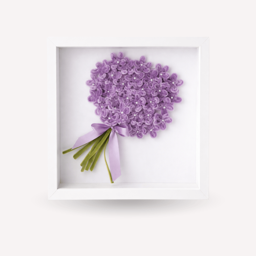 Quadro ricordo artigianale con cornice bianca, bouquet di fiori lilla fatti a mano con piccoli dettagli luminosi, fiocco lilla chiaro e steli verdi. Decorazione elegante e personalizzabile, adatta come idea regalo per la festa della donna o complemento d’arredo