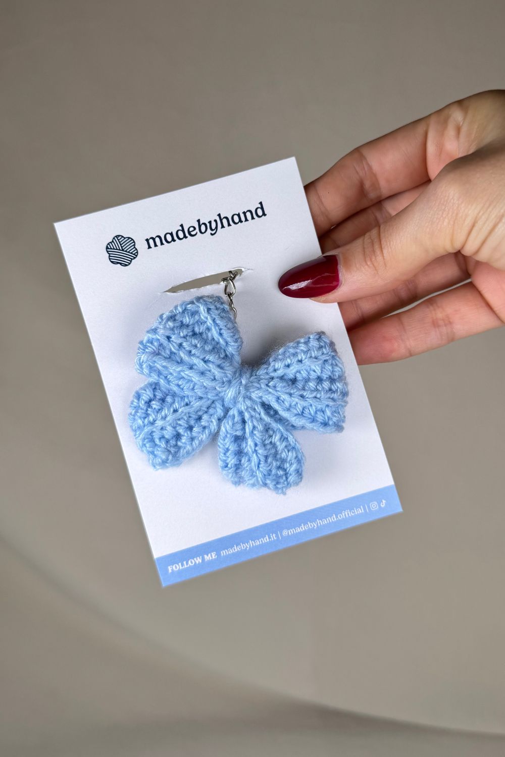 Portachiavi a fiocco azzurro realizzato all’uncinetto, fatto a mano in Italia da Made By Hand, presentato su cartoncino bianco con logo, ideale come bomboniera o regalo artigianale sostenibile