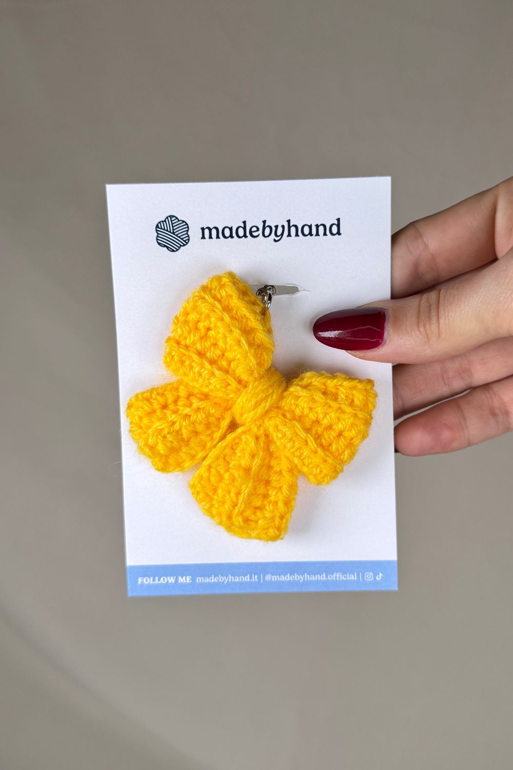 Portachiavi a fiocco giallo realizzato all’uncinetto, fatto a mano in Italia da Made By Hand, presentato su cartoncino bianco con logo, ideale come bomboniera o regalo artigianale sostenibile