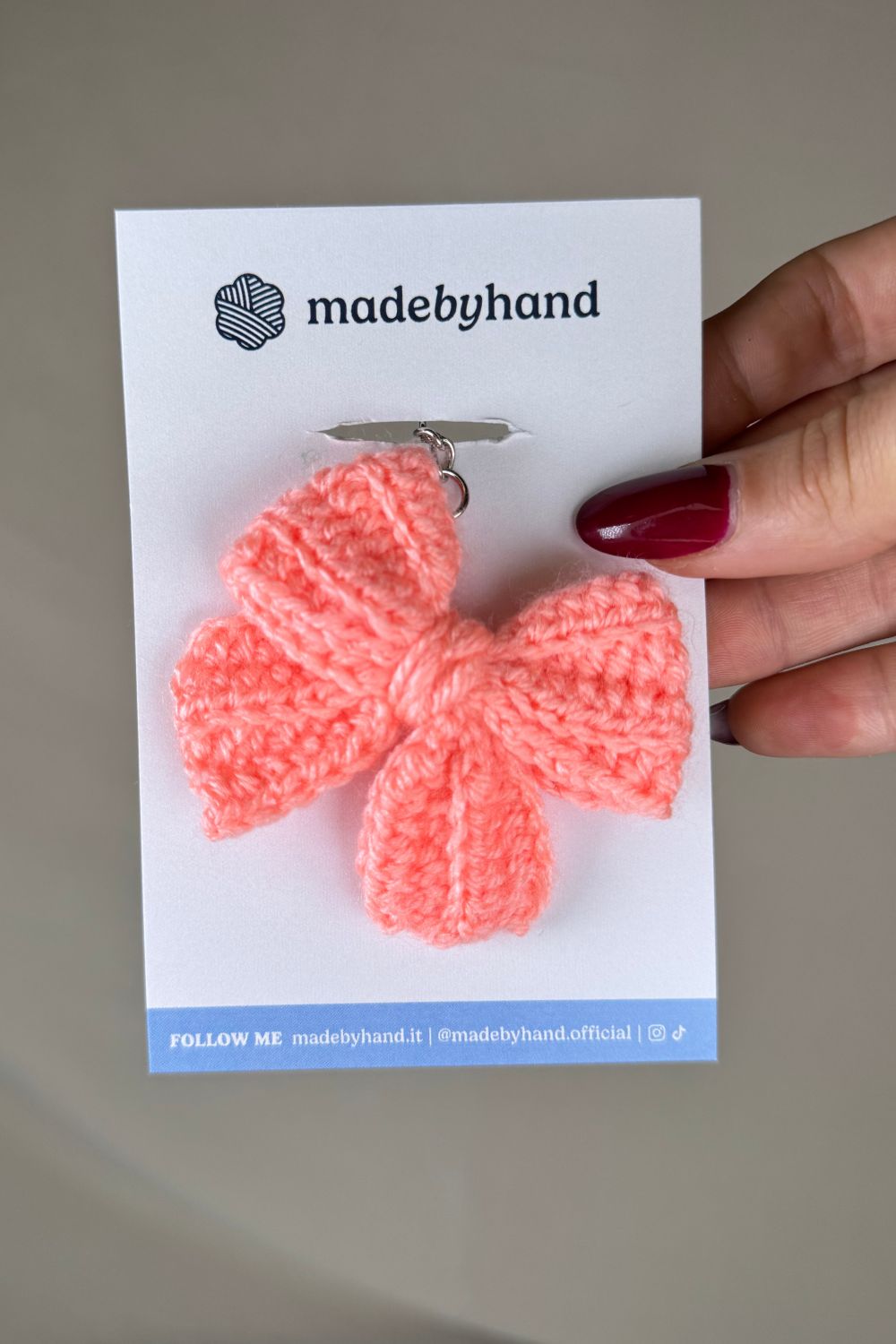 Portachiavi a fiocco rosa salmone realizzato all’uncinetto, fatto a mano in Italia da Made By Hand, presentato su cartoncino bianco con logo, ideale come bomboniera o regalo artigianale sostenibile