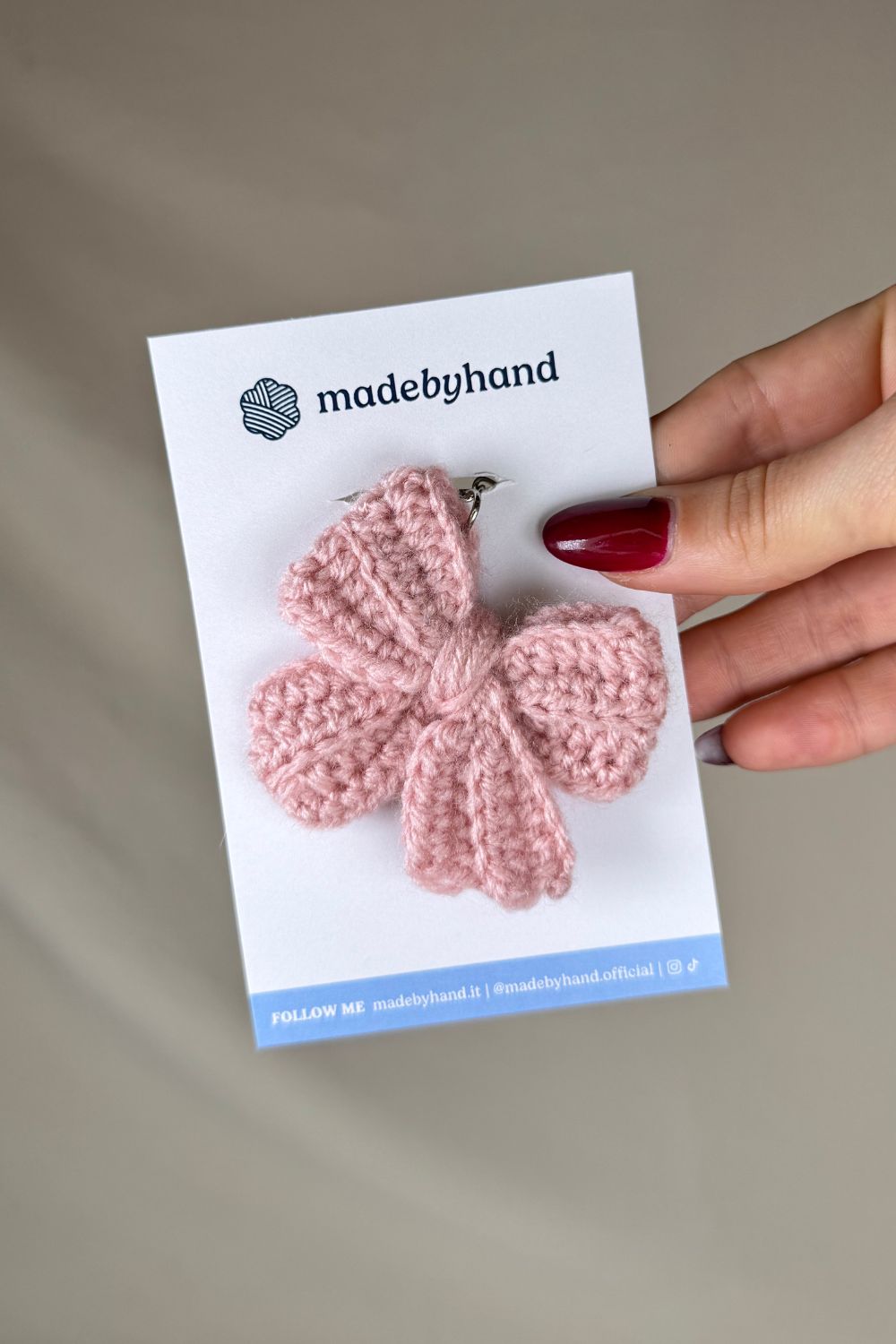 Portachiavi a fiocco rosa realizzato all’uncinetto, fatto a mano in Italia da Made By Hand, presentato su cartoncino bianco con logo, ideale come bomboniera o regalo artigianale sostenibile