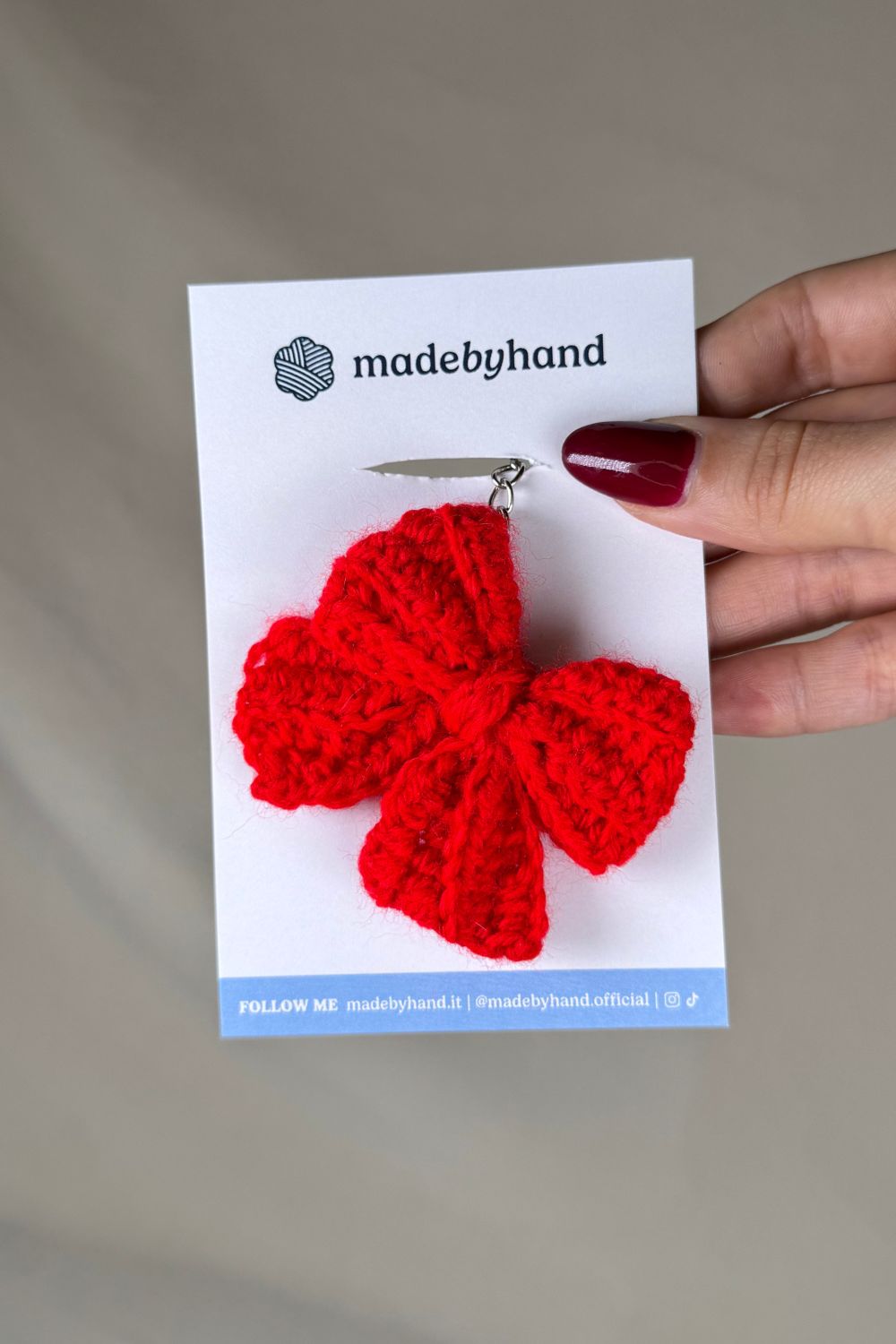 Portachiavi a fiocco rosso realizzato all’uncinetto, fatto a mano in Italia da Made By Hand, presentato su cartoncino bianco con logo, ideale come bomboniera o regalo artigianale sostenibile