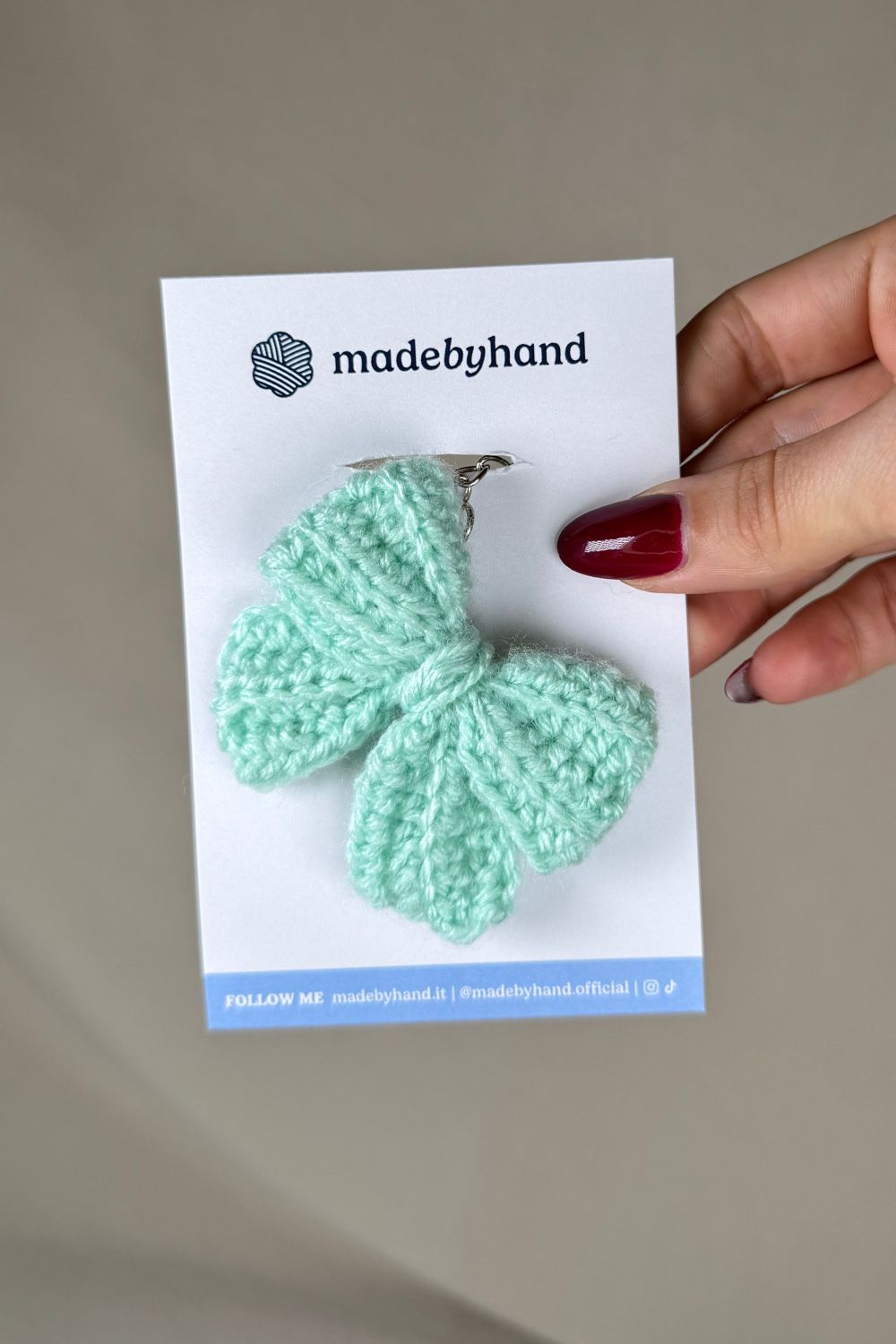 Portachiavi a fiocco verde tiffany realizzato all’uncinetto, fatto a mano in Italia da Made By Hand, presentato su cartoncino bianco con logo, ideale come bomboniera o regalo artigianale sostenibile