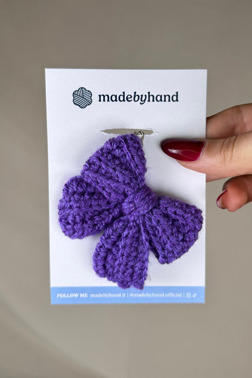 Portachiavi a fiocco viola realizzato all’uncinetto, fatto a mano in Italia da Made By Hand, presentato su cartoncino bianco con logo, ideale come bomboniera o regalo artigianale sostenibile
